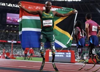 Tokyo 2020 Paralympic Games - Athletics - Men's 200m - T61 Final - Olympic Stadium, Tokyo, Japan - September 3, 2021. Ntando Mahlangu of South Africa celebrates after winning gold with the flag of South Africa whilst Regas Woods Sr of the United States and Luis Puertas of the United States look dejected REUTERS/Thomas Peter