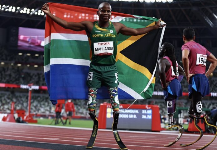 Tokyo 2020 Paralympic Games - Athletics - Men's 200m - T61 Final - Olympic Stadium, Tokyo, Japan - September 3, 2021. Ntando Mahlangu of South Africa celebrates after winning gold with the flag of South Africa whilst Regas Woods Sr of the United States and Luis Puertas of the United States look dejected REUTERS/Thomas Peter Tokyo 2020 Paralympic Games - Athletics - Men's 200m - T61 Final - Olympic Stadium, Tokyo, Japan - September 3, 2021. Ntando Mahlangu of South Africa celebrates after winning gold with the flag of South Africa whilst Regas Woods Sr of the United States and Luis Puertas of the United States look dejected REUTERS/Thomas Peter