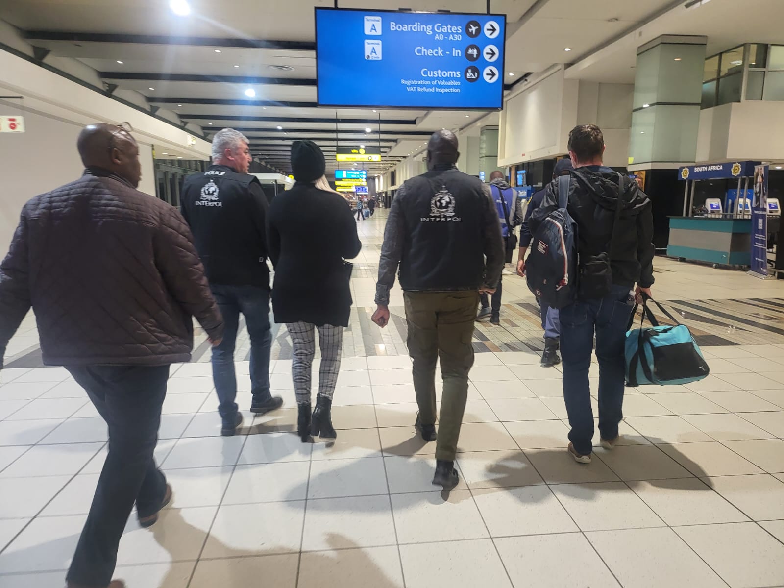 Wanted Ruth Lawrence with her blonde hair showing beneath her hat is escorted through Johannesburg Airport by two Interpol officers and one of the team of Irish detectives on the right carrying luggage heading for a waiting British Airways flight to Heathrow Airport.