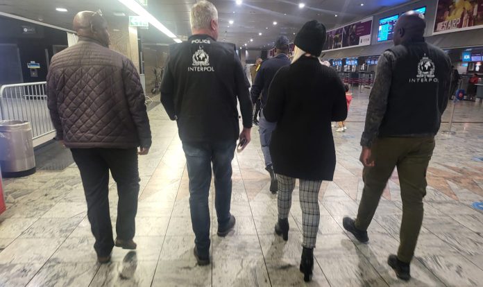 Wanted Ruth Lawrence with her blonde hair showing beneath her hat is escorted through Johannesburg Airport by two Interpol officers and one of the team of Irish detectives on the right carrying luggage heading for a waiting British Airways flight to Heathrow Airport.