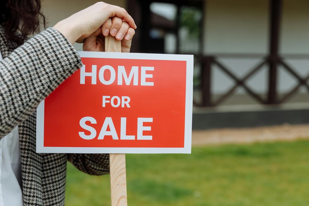 Real estate agent standing beside a "home for sale" sign. This image accompanies an article titled 'Residential property prices: Good news for South African homeowners'